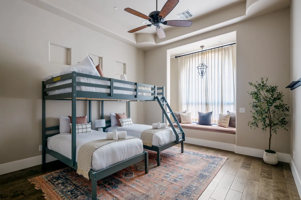 A playful and cozy guest bedroom with a bunk bed, window seat, area rug, and ceiling fan. The inviting space is perfect for kids or families, offering both a fun sleepover vibe and a quiet reading nook.