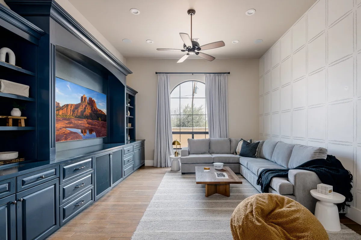 A contemporary media room setup with a large TV displaying a scenic landscape, a light-colored sectional sofa arranged for optimal viewing, a wooden coffee table, and decorative wall shelving. The space is designed for movie nights or relaxed gatherings with friends and family.