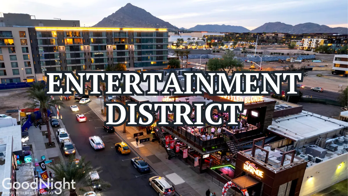 A lively street view of Scottsdale's Entertainment District, featuring prominent signage, bustling restaurants and bars, and a backdrop of scenic mountains. Cars line the street, and the energy of nightlife is palpable.