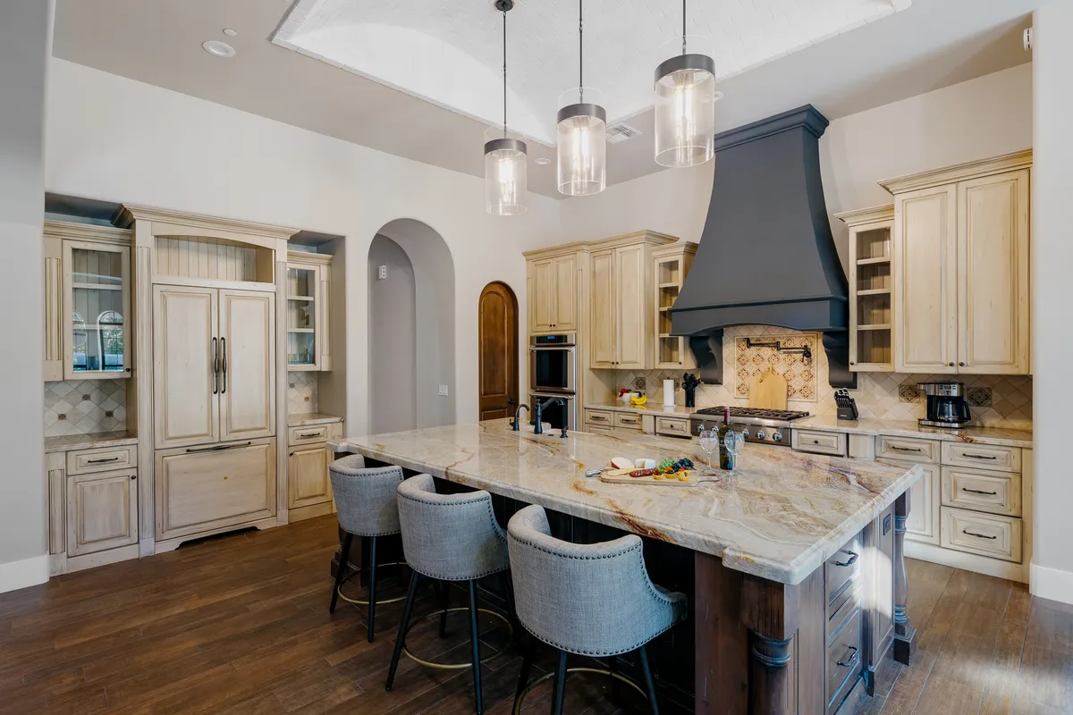 A spacious, welcoming kitchen featuring a large island perfect for meal prep and gathering, illuminated by elegant chandelier lighting. The decorative hood over the stove and rich wooden cabinetry add a touch of luxury and warmth, making the space ideal for both cooking and entertaining.