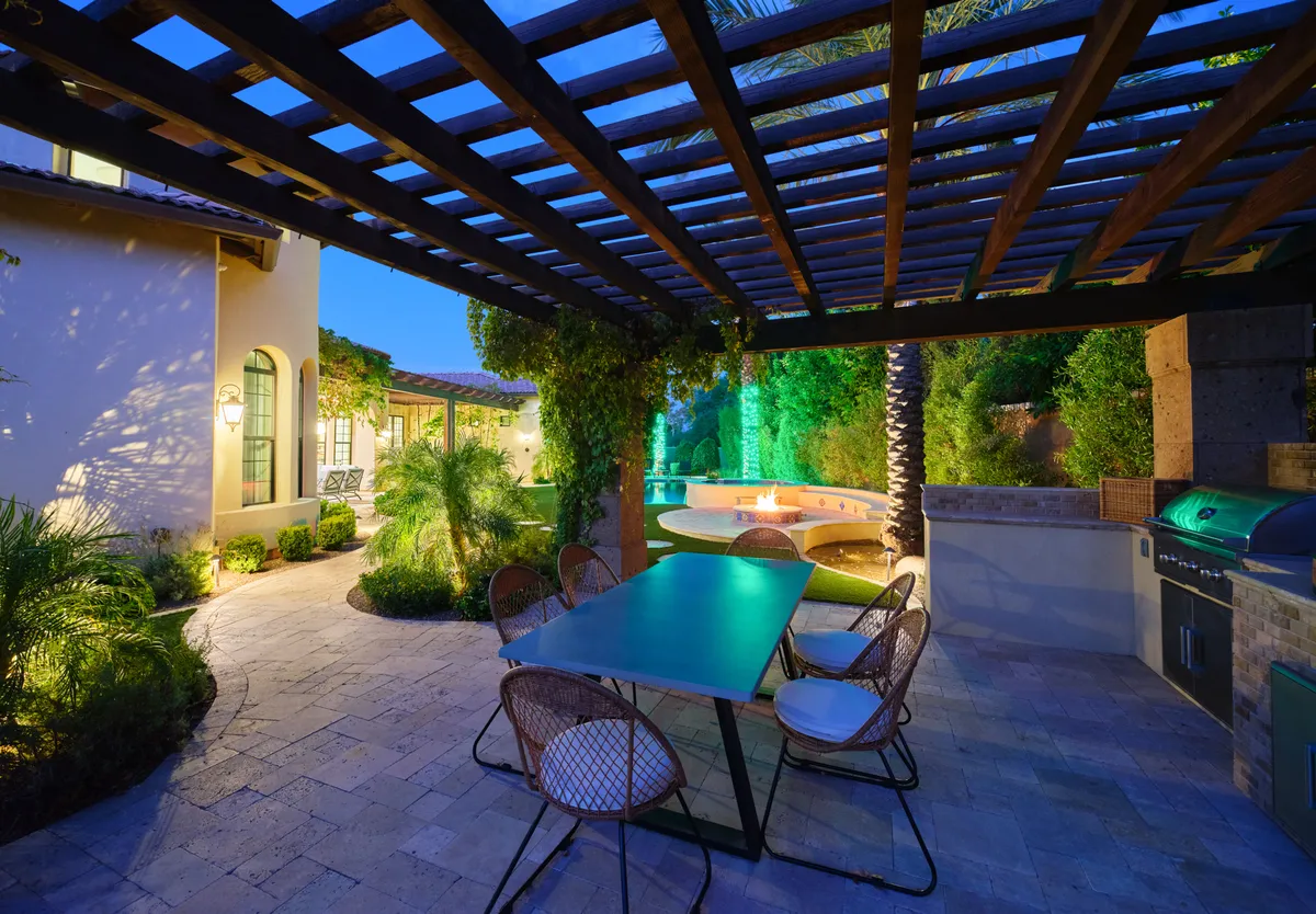 A beautifully arranged outdoor dining space under a pergola, with a stylish dining table set next to the swimming pool. The area also includes an outdoor grill, lush greenery, and ambient lighting perfect for evening gatherings or alfresco meals.