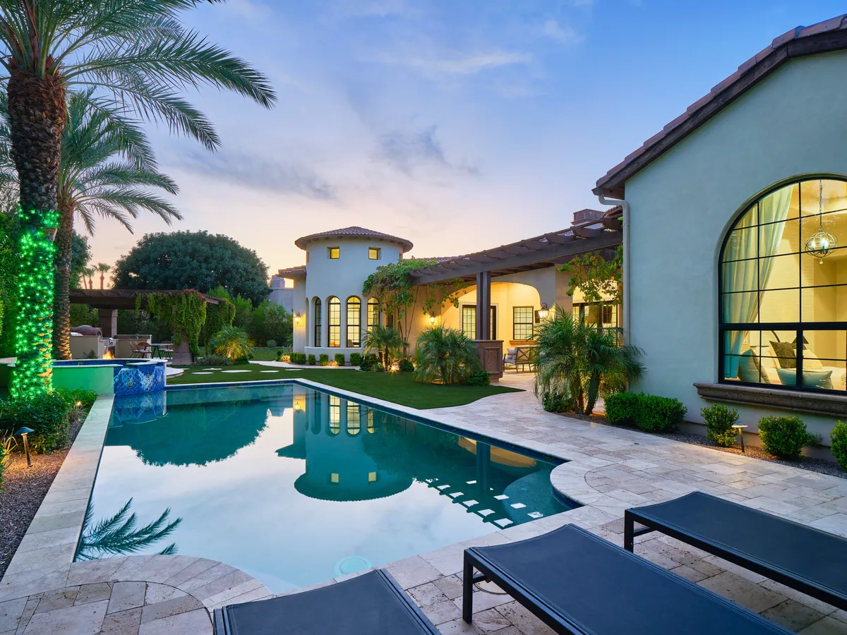 The image showcases a stunning Scottsdale estate exterior at dusk, with a shimmering swimming pool surrounded by elegant lounge chairs and lush palm trees. The home's modern architecture is highlighted by soft, ambient lighting, inviting guests to relax poolside or gather for evening festivities under the Arizona sky.
