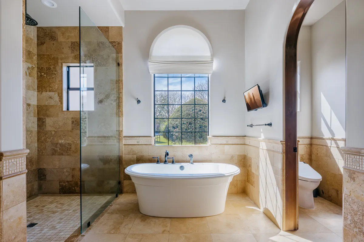 A luxurious en-suite bathroom featuring a freestanding bathtub by a large window, a spacious shower area, and a TV mounted on the wall. The space is filled with natural light, exuding a spa-like atmosphere for ultimate relaxation.