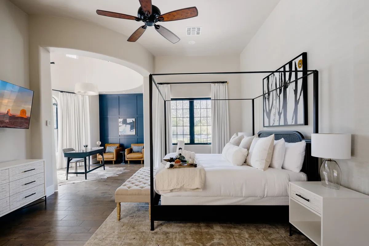 A tranquil and sophisticated bedroom featuring a four-poster bed with plush bedding, a cozy seating area for relaxing or reading, elegant artwork on the wall, and a TV for personal entertainment. The room exudes comfort and privacy, perfect for a restful night's sleep.