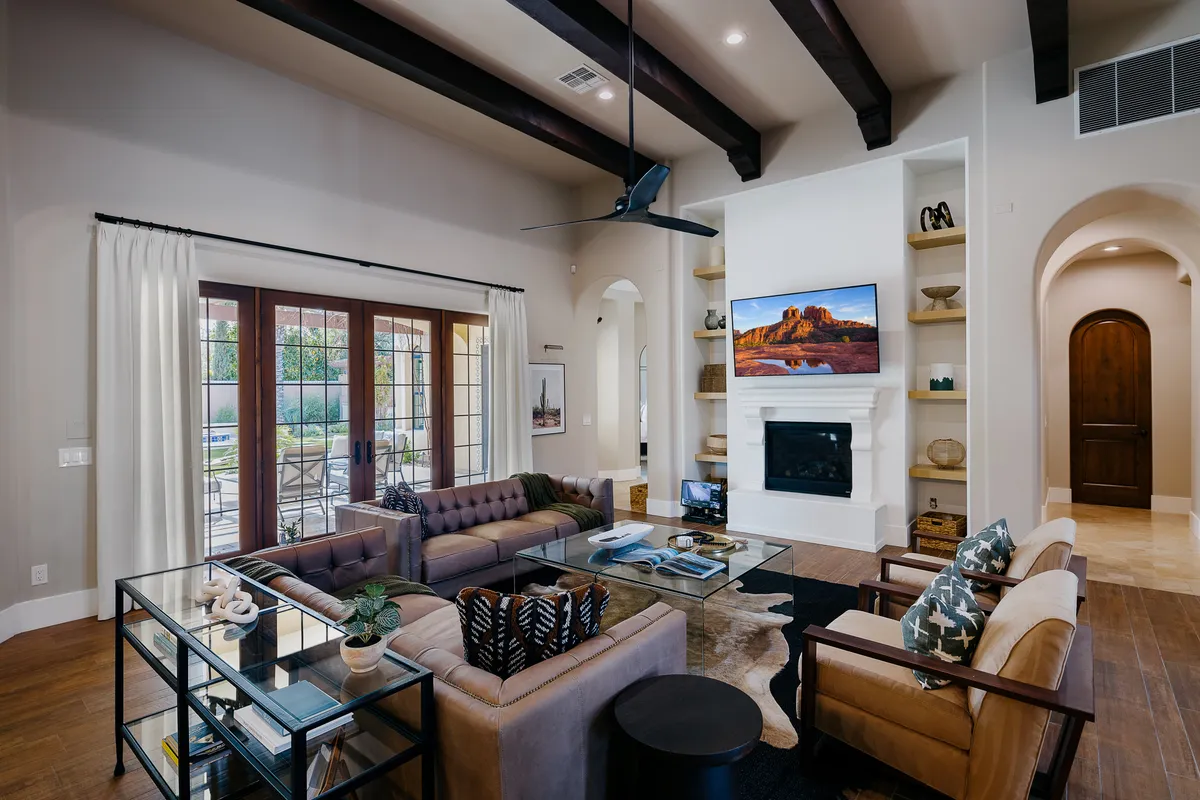 A warmly lit living room featuring a plush chocolate brown tufted sofa, a glass coffee table, a flat-screen TV mounted above a modern fireplace, and expansive windows with flowing white curtains. Neutral decorative shelves and ambient sunlight create a cozy, welcoming setting ideal for relaxing or entertaining.