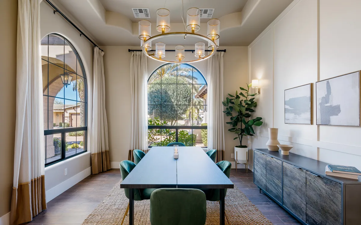 An elegant dining area featuring a sophisticated table set beneath a sparkling chandelier. Large windows bathe the room in sunlight, while greenery and artistic touches create a refined yet inviting atmosphere for any meal.