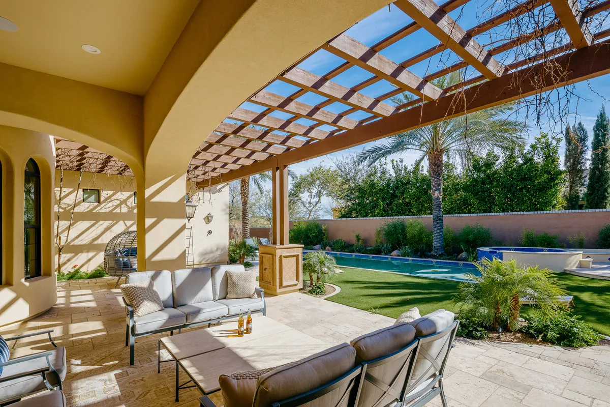 A wide view of the backyard pool area featuring a sparkling swimming pool, comfortable outdoor seating under a pergola, lush landscaping with palm trees and greenery, and space for sunbathing or entertaining guests. The patio area is bathed in warm sunset light, creating a relaxed and inviting atmosphere.